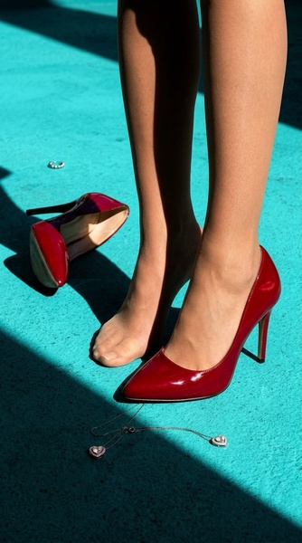 A close-up vertical shot focusing on red patent leather high heels and discarded jewelry on a vibrant teal background, conveying luxury and spontaneity.