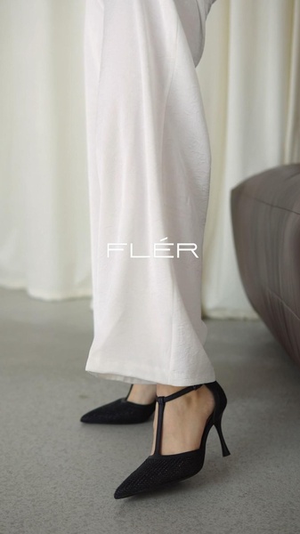 A close-up aesthetic shot of black stiletto high heels worn with white wide-leg trousers, emphasizing clean lines and a premium brand identity.