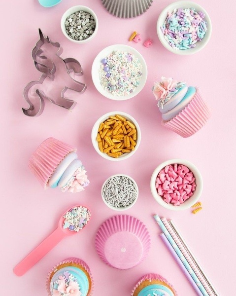 A high-angle flat lay photograph of cupcake baking supplies including a unicorn-shaped cookie cutter, sprinkles, and finished cupcakes in pastel colors on a soft pink background.
