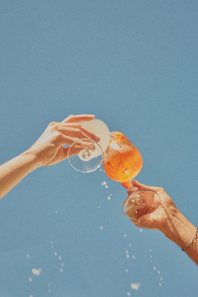 A close-up, low-angle shot of two people toasting with orange-hued cocktails against a clear, bright blue summer sky.