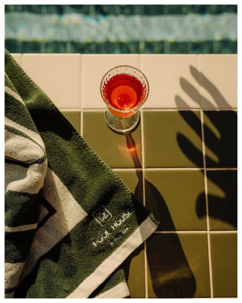 A close-up, high-contrast shot of a glass of red beverage resting on a tiled pool deck next to a branded pool towel, using natural sunlight and strong shadows.