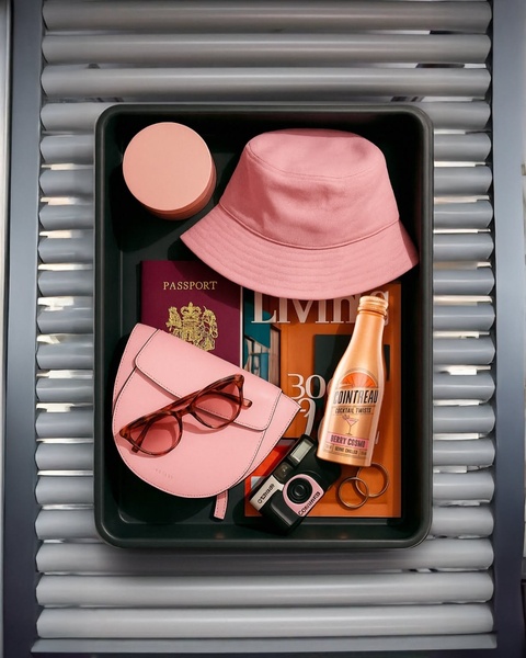 A high-end overhead photograph featuring a curated collection of travel essentials including a cocktail bottle, hat, and sunglasses.