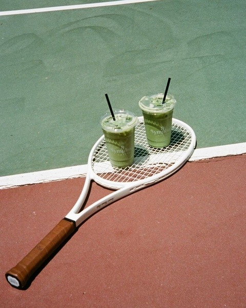 A high-angle photograph showing two iced matcha latte cups resting on a tennis racket placed on a court, emphasizing a leisure lifestyle.