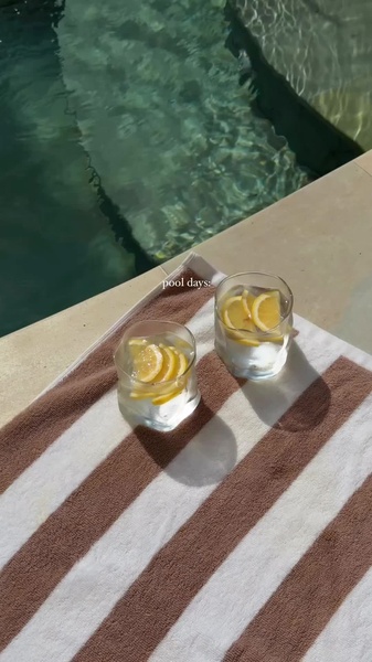 A high-quality 9:16 vertical shot of two glasses of water with lemon slices sitting on a striped towel by a pool, capturing a serene summer mood.