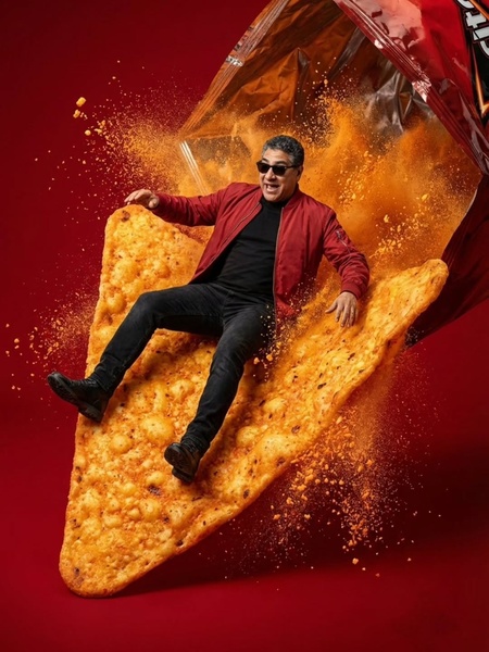 A man in sunglasses and a red jacket floats on a giant corn chip emerging from a bag, surrounded by an explosion of seasoning.