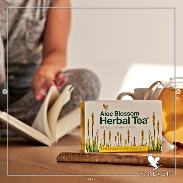A close-up, warm-toned composition featuring a box of herbal tea next to a glass of brewed tea with a blurred person reading a book in the background.