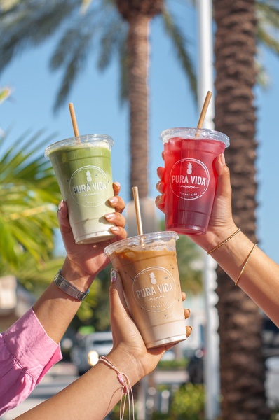 Three hands holding different refreshing drinks against a sunny palm tree backdrop to showcase variety and social connection.