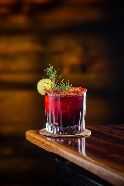 A high-contrast, moody close-up of a cocktail in a rocks glass, garnished with rosemary and lime, set against a dark, warm background.