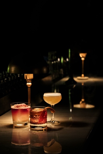 A dark, atmospheric photograph of three curated cocktails on a marble bar top, illuminated by a single warm desk lamp.