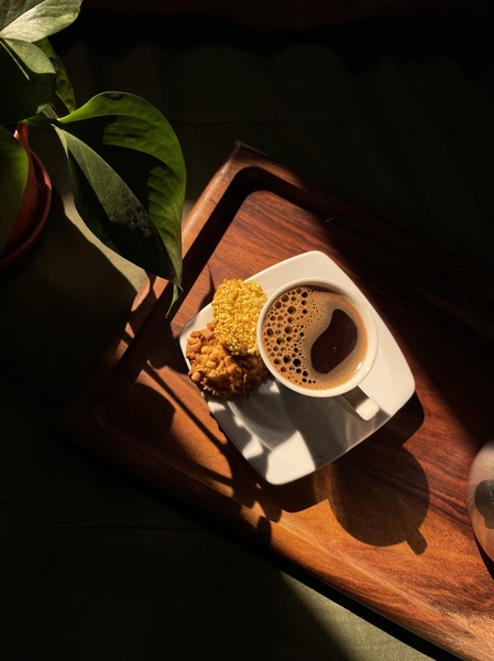 A high-angle, top-down shot of a cup of black coffee and two cookies on a wooden tray, set in soft, natural morning light.