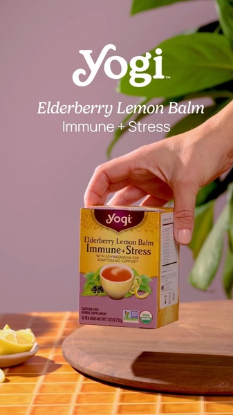 A close-up shot of a human hand holding a box of herbal tea against a minimalist, purple-toned background with natural greenery.
