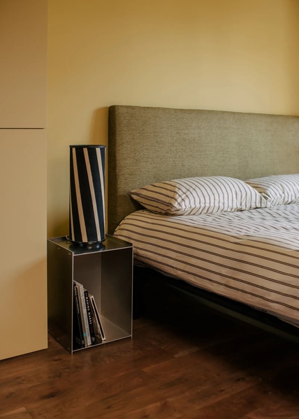 A clean, lifestyle-focused bedroom shot featuring a modern metal bedside table, a patterned lamp, and a warm color palette.