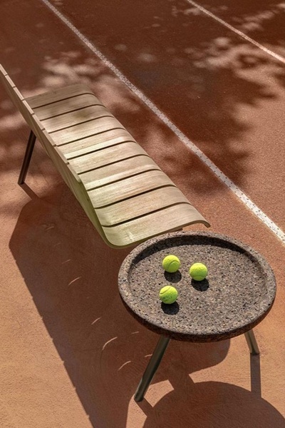 A high-angle shot showcasing a wooden garden bench and a circular pebble-texture side table with three tennis balls on a tennis court.