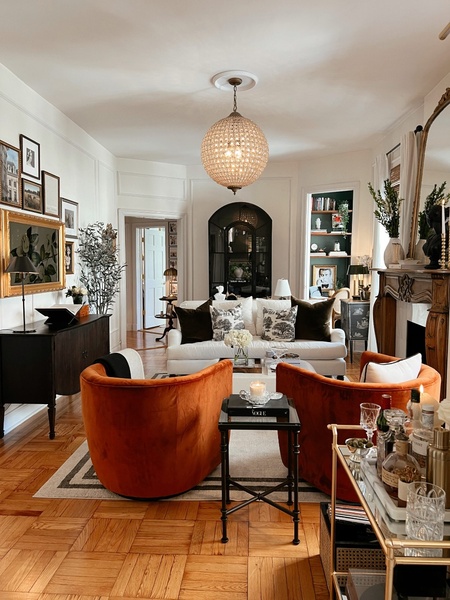 A high-end, professionally styled living room featuring deep orange velvet chairs, a central crystal chandelier, and a sophisticated, layered interior design aesthetic.