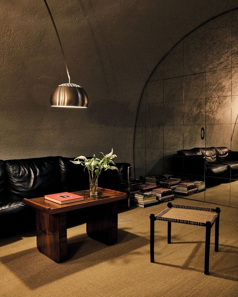 A high-end, dark-toned interior scene featuring a minimalist wooden-weave stool as the focal point within a vault-like, moody living space.