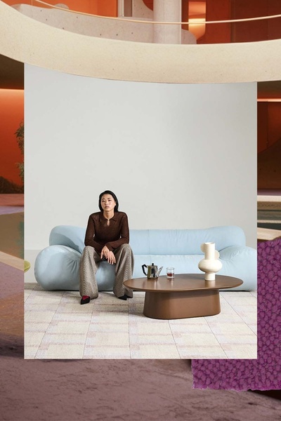 A high-end studio shot of a person sitting on a light blue curved sofa, accented by a neutral rug, a coffee table, and an artistic vase.