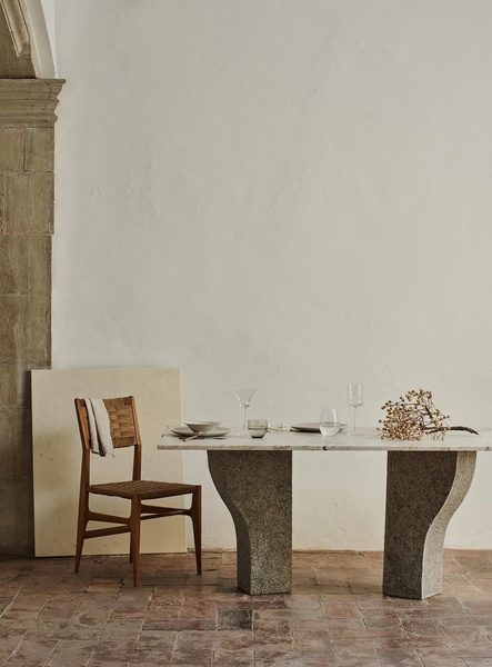 A high-end, editorial-style product shot featuring a natural stone dining table and a woven chair against a textured white wall.
