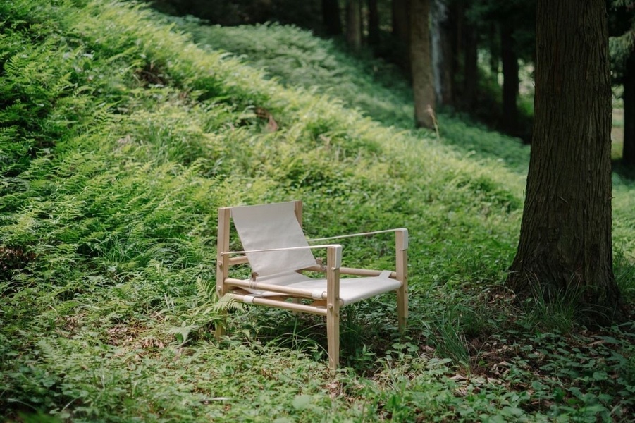 A high-quality product shot of a simple, modern folding chair placed deep in a lush, green forest setting.