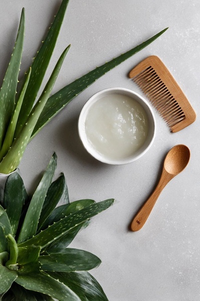 A top-down, minimalist product-hero shot showcasing raw Aloe Vera leaves, a wooden comb, and a bowl of prepared gel on a neutral grey background.
