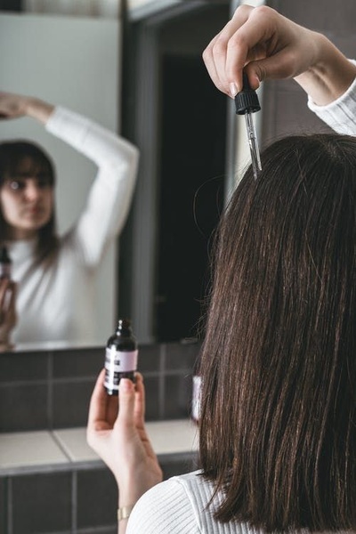 A close-up, first-person perspective of a consumer applying a scalp serum directly to their roots in a bathroom mirror setting.