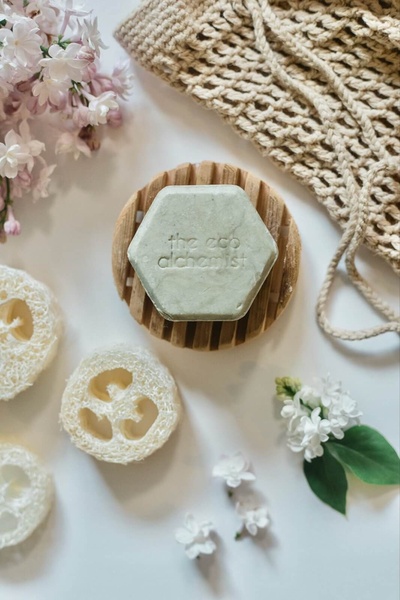 This creative features a solid shampoo bar centered on a wooden soap dish, surrounded by natural elements like loofah sponges and fresh flowers to emphasize an organic aesthetic.