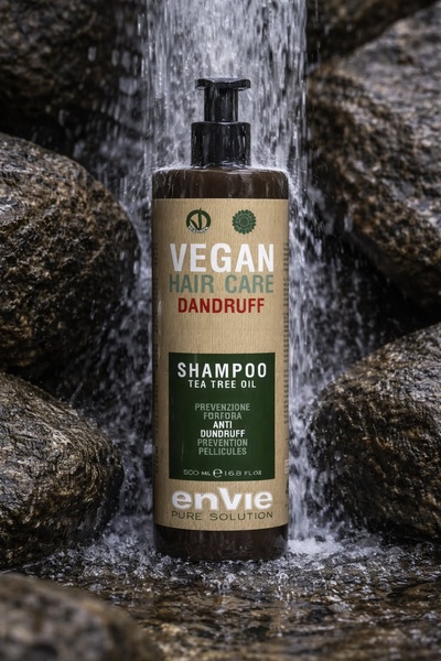 A high-contrast, organic product photography shot featuring a vegan dandruff shampoo bottle positioned directly under a cascading waterfall onto natural stone.