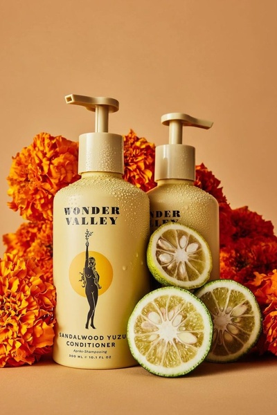 A high-fidelity studio shot featuring two conditioner bottles surrounded by marigold flowers and fresh yuzu slices, utilizing a warm, monochromatic color palette.