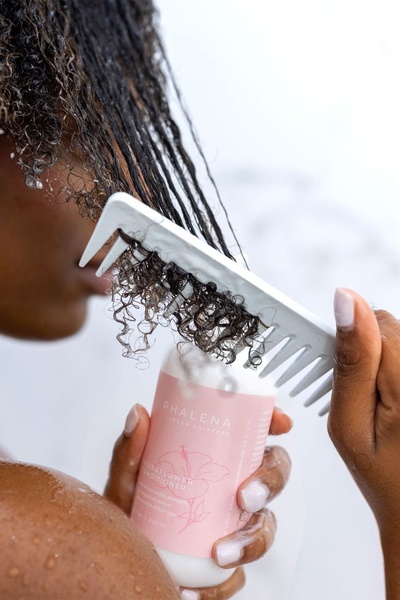 A close-up, sensory-focused shot showing conditioner being combed through curly hair in the shower to demonstrate product texture and performance.