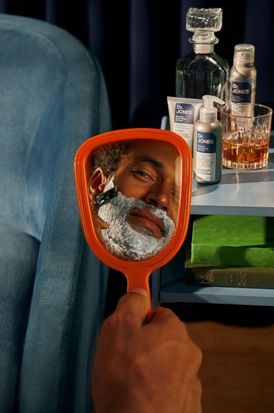 A man's face being shaved is captured through a hand mirror, highlighting the grooming routine against a moody, sophisticated background.