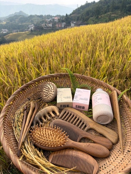 A flat-lay photograph of rice water hair products and wooden combs arranged in a basket against a backdrop of a terraced rice field.