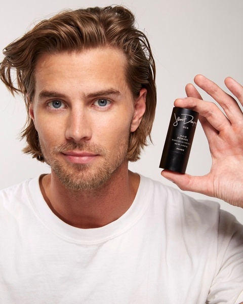 A close-up portrait of a model holding a hair pomade product, utilizing a minimalist, high-key studio aesthetic to drive brand trust.
