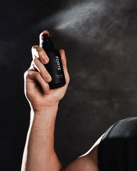 A close-up shot capturing a hand actively spraying a matte black bottle, with a fine mist visible against a dark, moody background.