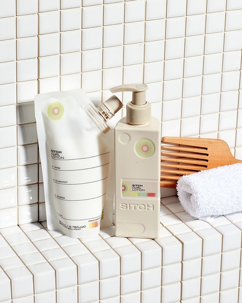 A high-quality product photography shot featuring a curl lotion bottle and its matching refill pouch on a clean tiled bathroom backdrop.