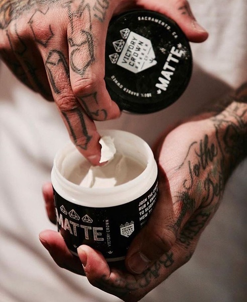 A close-up, high-contrast shot of a hand with tattoos scooping a small portion of matte hair pomade from its container.