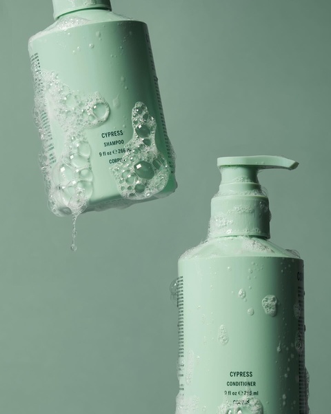 A clean, studio-lit shot of shampoo and conditioner bottles suspended in mid-air surrounded by fresh soap bubbles on a solid sage green background.
