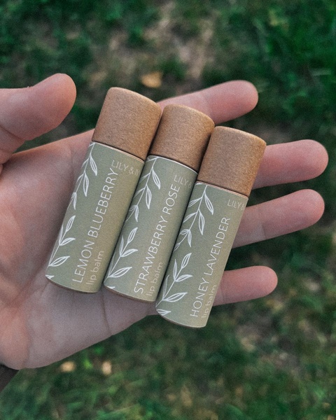 A close-up, top-down shot of a hand holding three different natural lip balm tubes against a blurred outdoor background.