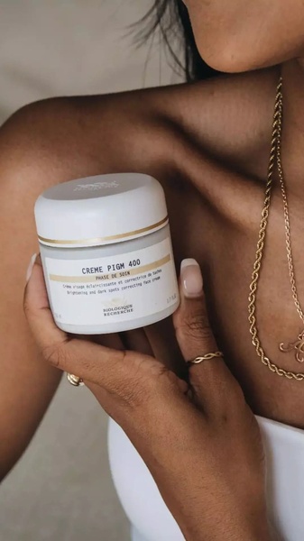 A close-up aesthetic shot of a jar of face cream held by a person, focusing on the product packaging in a clean, high-end style.