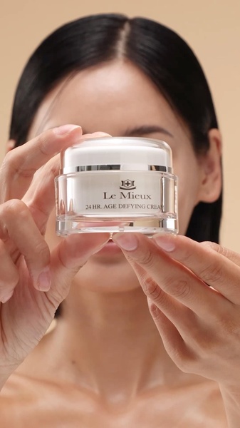 A close-up, high-quality shot of a woman holding a jar of age defying cream, focusing on the product packaging against a neutral, warm background.