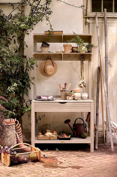 A high-quality lifestyle shot featuring a wooden garden potting bench styled with rustic tools, clay pots, and fresh produce against a neutral outdoor wall.