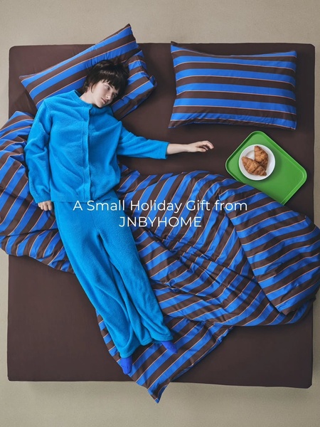 A top-down, cozy lifestyle shot featuring a person in blue fleece pajamas lying on striped bedding with a small breakfast tray.