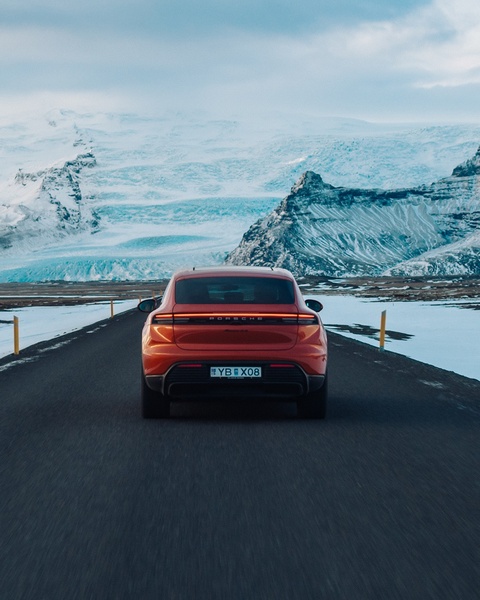 A high-contrast rear view of a luxury electric sports car driving on a coastal road framed by a dramatic, snow-capped mountain range.