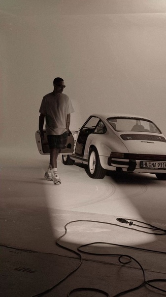 A high-contrast, sepia-toned shot of a person walking away from a vintage Porsche 911. The composition emphasizes a premium, cinematic lifestyle aesthetic.