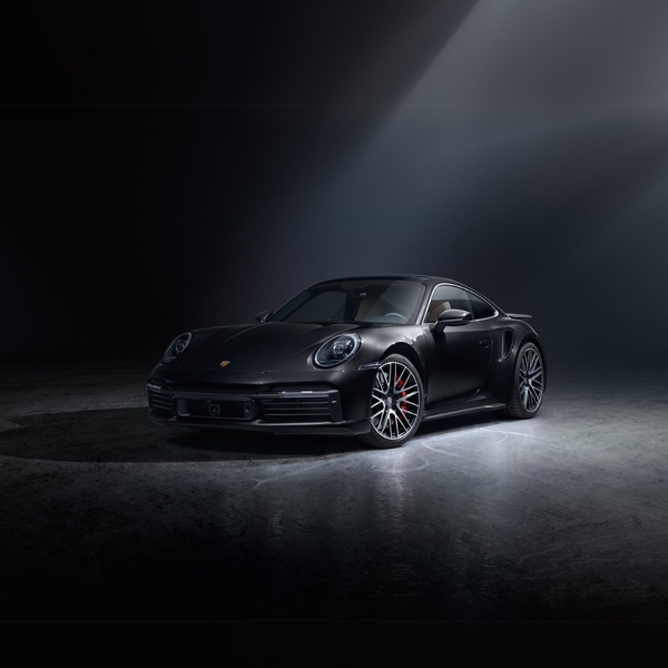 A sleek, black luxury sports car positioned in a minimalist, dimly lit studio environment with dramatic top-down lighting.