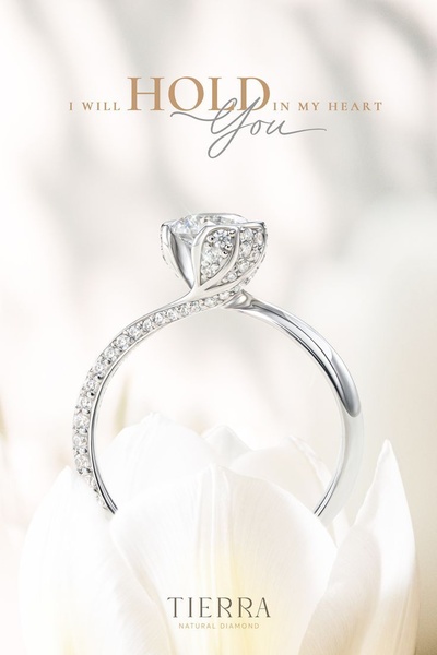 A refined, close-up hero shot of a diamond engagement ring resting on a white flower petal, focusing on clean aesthetics and emotional copy.