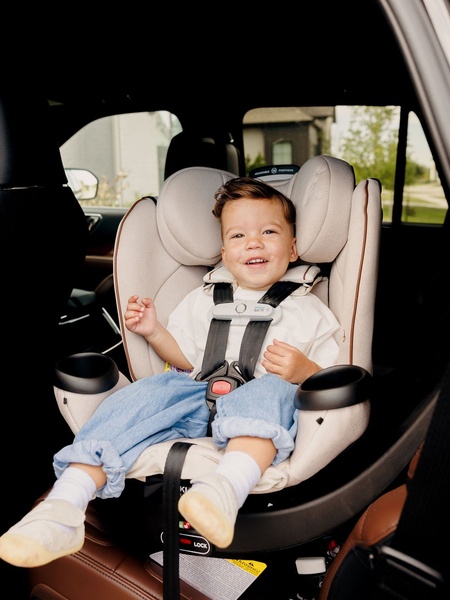 A high-quality, lifestyle-oriented shot featuring a smiling toddler securely fastened in a beige car seat within a car.