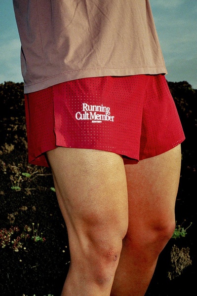 A close-up, high-contrast shot of red performance shorts on a runner, highlighting the 'Running Cult Member' branding in a natural, outdoor setting.