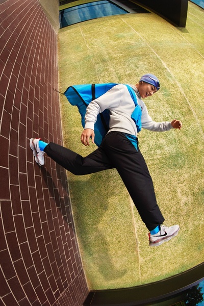A high-angle, wide-lens shot of a person mid-air wearing athletic gear and running shoes, captured against a brick wall and grassy ground.