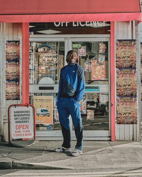 A man stands confidently in front of an off-license shop wearing a modern blue and black tracksuit, utilizing natural lighting and real-world placement.