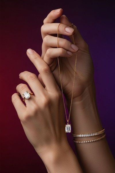 A close-up photograph featuring a hand wearing an emerald-cut ring while holding a matching necklace, presented against a deep gradient background.