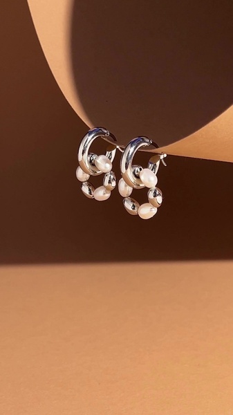 A close-up studio shot of silver hoop earrings decorated with small pearls, captured against a clean, warm-toned curved background.